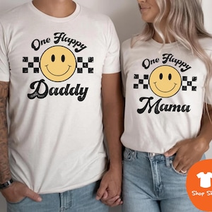 May include: Two off-white short-sleeve t-shirts featuring a yellow smiley face graphic with black checkered patterns on the sides. The left shirt displays the text One Flappy Daddy, while the right shirt displays the text One Flappy Mama in black.