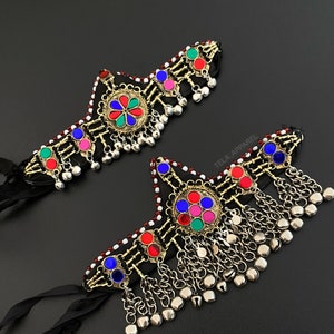 May include: Two decorative headbands with vibrant jewel embellishments and hanging silver-coloured charms. The headbands have a black fabric base, detailed metalwork, and a central floral design with red, blue, green, and pink gems. Black ribbons extend from the sides.