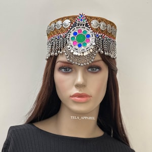 Peut inclure: Un bandeau brun et or avec des embellissements argentés et un grand pendentif circulaire avec des pierres colorées. Le bandeau est orné d'une frange de chaînes et de clochettes argentées.