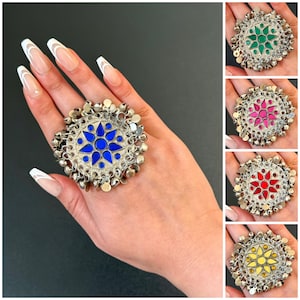 Può includere: Un grande anello a dichiarazione argentato con un design centrale a stella blu. L'anello è circondato da piccole campanelle pendenti. Vengono mostrati anche anelli aggiuntivi con centri verdi, rosa, rossi e gialli.