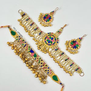 Può includere: Un set di gioielli dorati, tra cui una collana, due orecchini e un copricapo. I pezzi sono adornati con pietre preziose colorate e piccole campane dorate. Il testo "TELA APPAREL" è visibile.