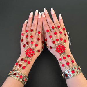 May include: A pair of hand jewelry featuring red circular accents and gold-toned chains. The jewelry includes finger rings connected to a central palm piece and a cuff bracelet. The jewelry is displayed against a black background.