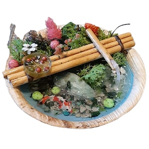 May include: A miniature zen garden with a small pond, a wooden bridge, and a waterfall. The pond is filled with blue resin and has small green lily pads and orange and white koi fish. The garden is surrounded by green moss, pink flowers, and a pine cone.