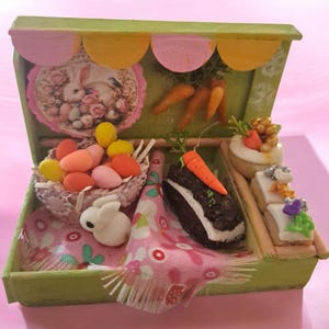 May include: A miniature Easter-themed display box with a green exterior. Inside, a basket of colorful eggs, a white bunny, and a chocolate cake with a carrot are visible. The backdrop features a bunny image and carrots.