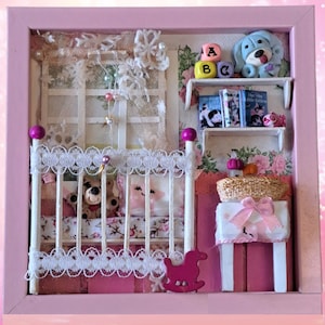 May include: A pink framed miniature nursery scene with a white crib, a rocking horse, a small table with a basket, and a shelf with books and a blue toy dog. The crib has a white lace trim and a pink and white floral patterned bedding. The rocking horse is pink and white. The table is white with a pink and white floral patterned tablecloth. The shelf is white with a pink and white floral patterned background. The books are colorful and have titles like "The Little Engine That Could" and "The Very Hungry Caterpillar". The toy dog is blue and white and has a red nose.