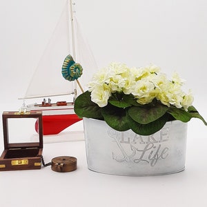 Puede incluir: Maceta de metal blanco con el texto "Lake Life" y un diseño de ancla. La maceta está llena de flores blancas y hojas verdes. Un pequeño modelo de velero está en el fondo.