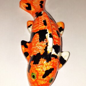 Puede incluir: Una peque&ntilde;a figurita de pez koi naranja y negro con ojos verdes. La figurita est&aacute; hecha de cer&aacute;mica y tiene una superficie texturizada.