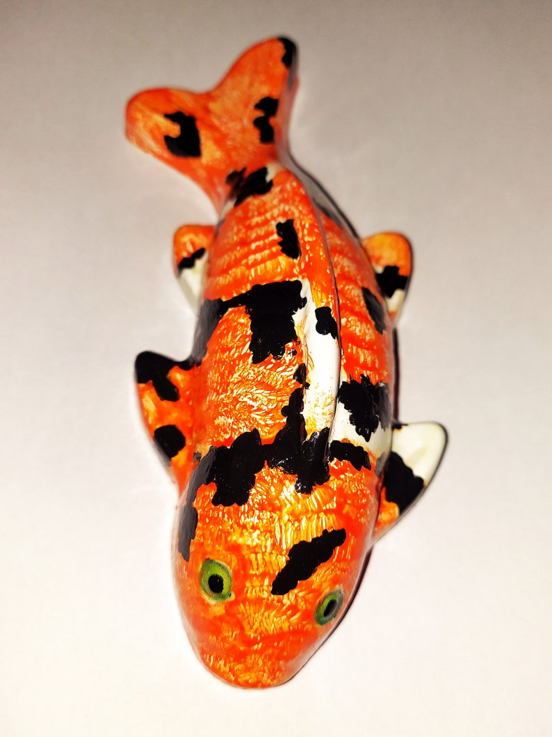 Puede incluir: Una peque&ntilde;a figurita de pez koi naranja y negro con ojos verdes. La figurita est&aacute; hecha de cer&aacute;mica y tiene una superficie texturizada.