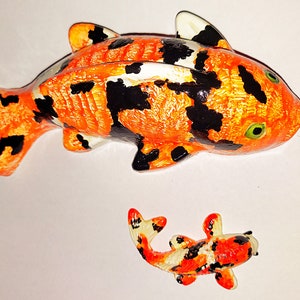 Puede incluir: Dos peces koi de cer&aacute;mica pintados a mano, uno grande y otro peque&ntilde;o. El pez m&aacute;s grande es naranja y negro con detalles blancos. El pez m&aacute;s peque&ntilde;o es naranja, negro y blanco.