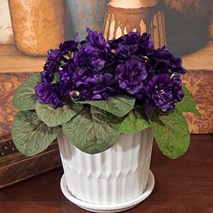May include: A white ceramic planter with a textured design holds a potted artificial violet plant with dark purple flowers and green leaves.