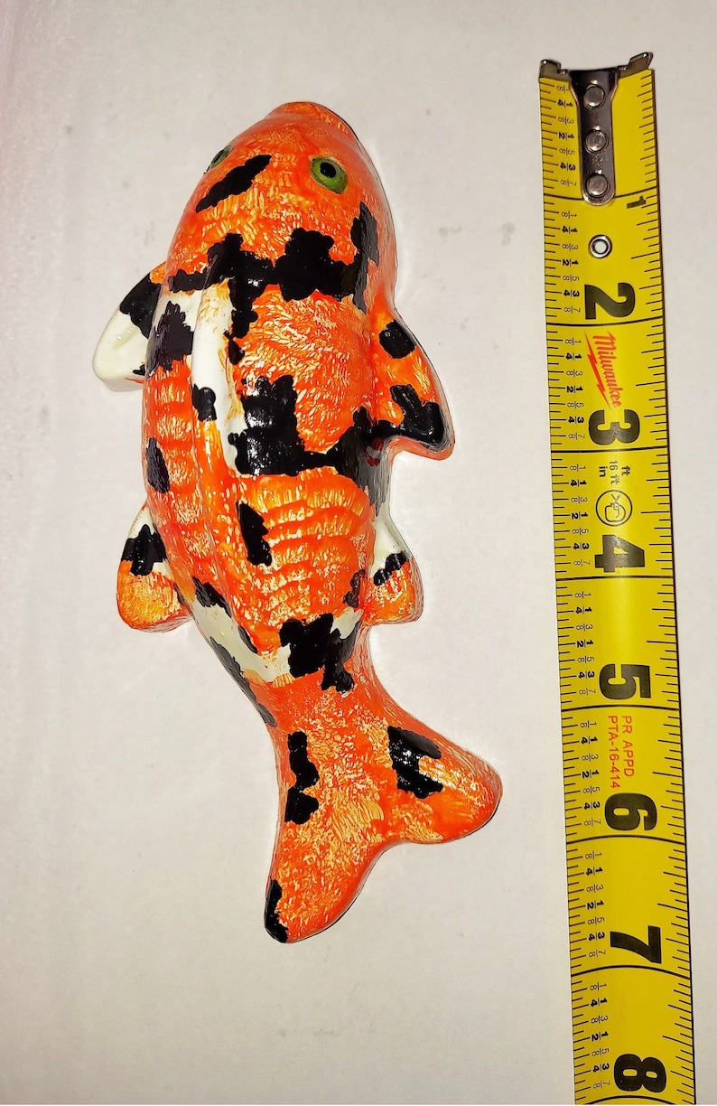 Puede incluir: Una figurita de pez koi pintada a mano en naranja, negro y blanco. La figurita mide aproximadamente 15 cm de largo y est&aacute; hecha de un material similar a la cer&aacute;mica.