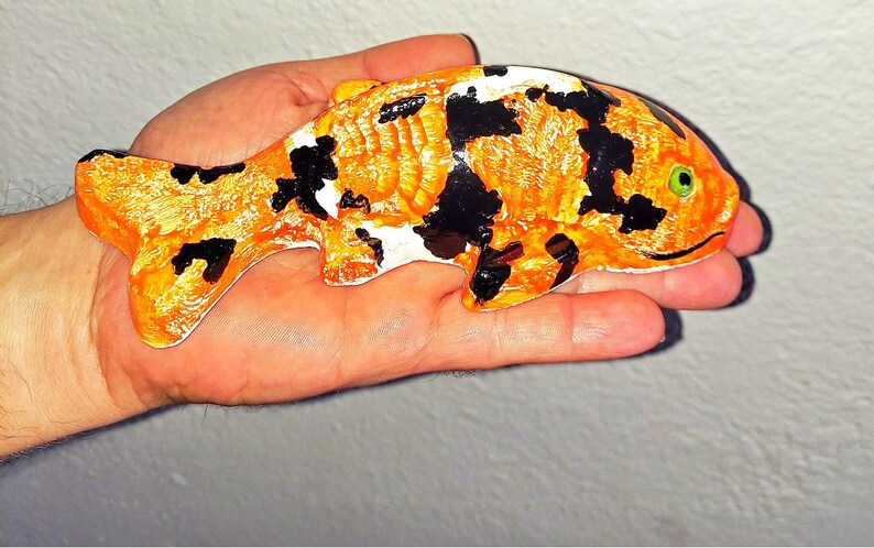 Puede incluir: Una figurita de cer&aacute;mica de pez koi pintada a mano con escamas naranja, negras y blancas. El pez tiene un ojo verde y una boca ligeramente hacia arriba.