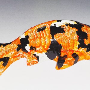 Puede incluir: Figurita de cer&aacute;mica pintada a mano de un pez koi con escamas naranja, negras y blancas. El pez tiene un ojo verde y la boca ligeramente abierta.