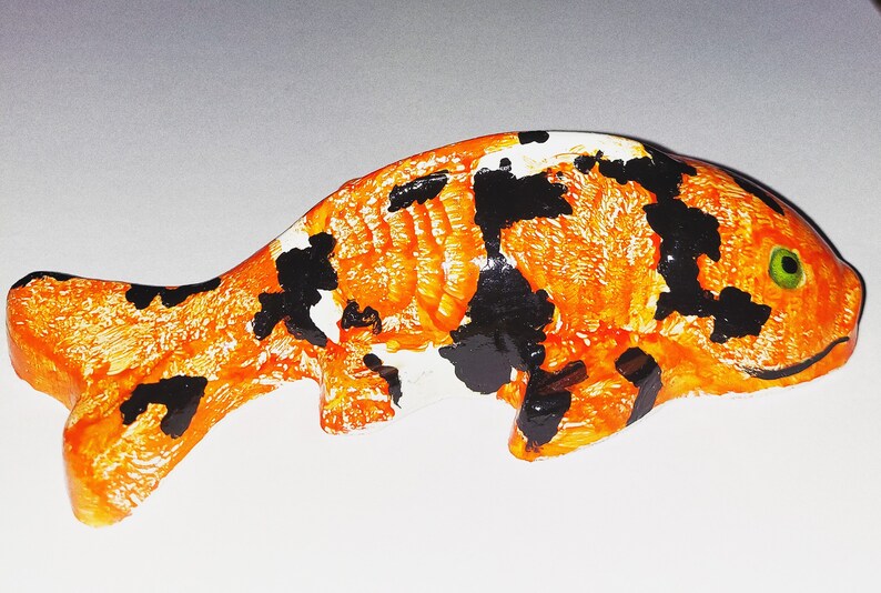 Puede incluir: Figurita de cer&aacute;mica pintada a mano de un pez koi con escamas naranja, negras y blancas. El pez tiene un ojo verde y la boca ligeramente abierta.