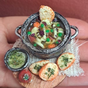 May include: A miniature food display featuring a dark, textured pot filled with soup containing noodles, green peas, and orange carrot slices, topped with a bread slice. Beside it sits a small bowl of green sauce, a red tomato, and three herb-topped bread slices.