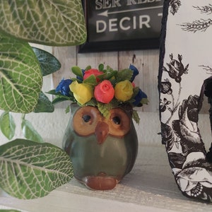 May include: A green ceramic owl planter with a flower crown made of red, yellow, and blue flowers. The owl planter is sitting on a white shelf.