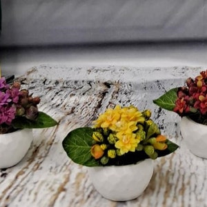May include: Three small white ceramic pots with different colored artificial flowers. The pots are on a whitewashed wooden surface. The flowers are purple, yellow, and red.