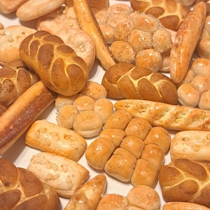 Puede incluir: Una variedad de panecillos y panes en miniatura, incluyendo una hogaza trenzada, una baguette y un panecillo redondo con semillas de sésamo. El pan es de color marrón claro y parece estar hecho de arcilla.