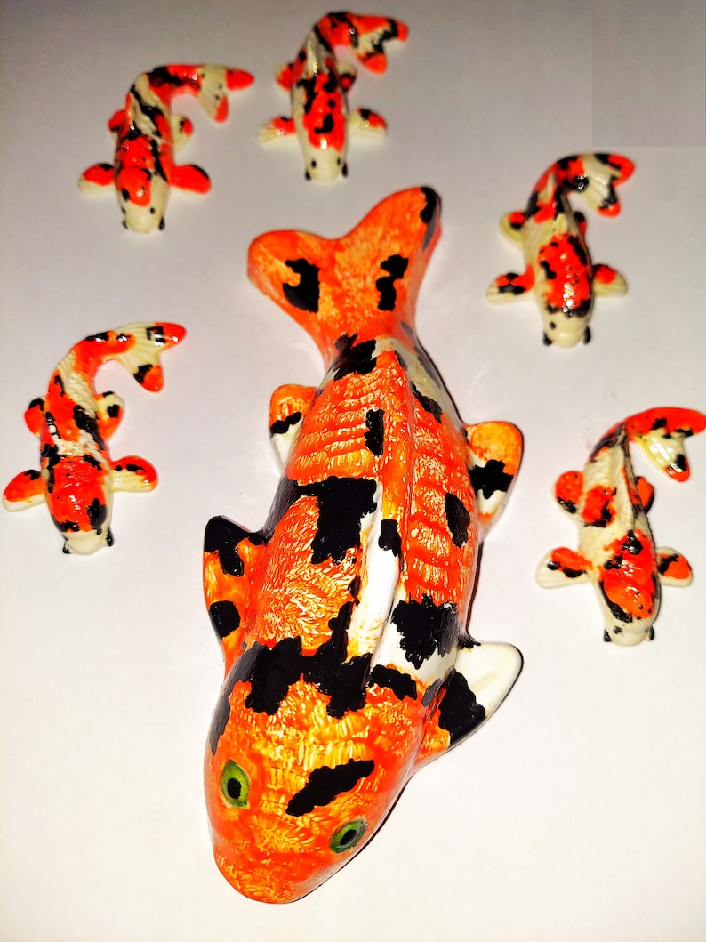Puede incluir: Cinco figuritas de peces koi de cer&aacute;mica en varios tama&ntilde;os. El pez m&aacute;s grande es naranja y negro con detalles blancos. Los peces m&aacute;s peque&ntilde;os son naranja, negro y blanco.
