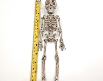 May include: A plastic skeleton with green eyes, measuring approximately 24 inches tall. The skeleton is light gray with visible rib cage, spine, and limbs. A yellow measuring tape is placed next to the skeleton.