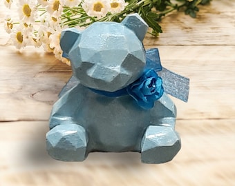 Puede incluir: Una figurita de oso geométrico azul claro con una cinta azul y un acento de flor azul. El oso está sentado sobre una superficie de madera clara.