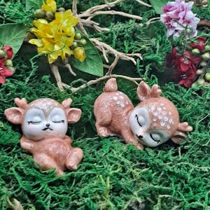 Puede incluir: Dos figuritas de ciervo de cerámica marrón con manchas blancas, tumbadas con los ojos cerrados, sobre un lecho de musgo verde.