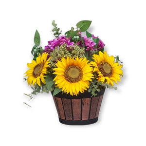 May include: A wooden planter with a rustic brown finish holds a bouquet of yellow sunflowers and pink flowers. The planter has a black base and is filled with green foliage.