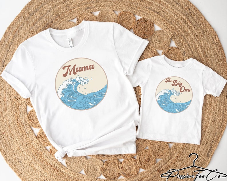 Op de afbeelding: Twee witte T-shirts met een blauwe golfgrafiek. Het grotere shirt zegt "Mama" in bruine tekst. Het kleinere shirt zegt "The Big One" in bruine tekst.