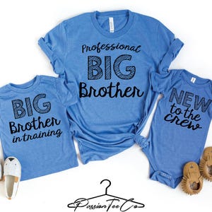 May include: Three blue shirts with black text that read "Professional Big Brother", "Big Brother in training", and "New to the crew". Two pairs of baby shoes and a pair of white and gray slip-on shoes are also in the image.