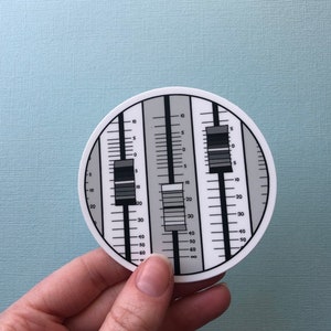 May include: A round sticker with a black, white, and gray design resembling a sound level meter. The sticker has three vertical columns with numbered scales and sliding bars. The sticker is held by a hand against a light blue background.