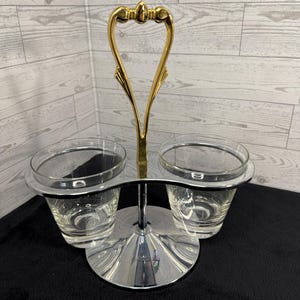 May include: A vintage two-part serving dish with a gold-colored handle and a chrome-plated base. The dish holds two clear glass cups. The handle has an ornate design. The base is round and reflective.