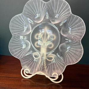 May include: A clear glass plate with a scalloped, flower-like edge and a central circular section. The plate features delicate, etched white lines radiating from the center to the outer rim, creating a shell-like pattern throughout the design.