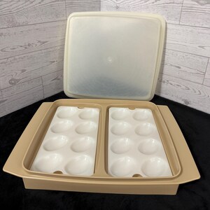 May include: A rectangular beige plastic egg storage container with a matching translucent lid. The base features two removable white plastic inserts, each containing eight molded oval indentations designed to hold sixteen eggs in total.