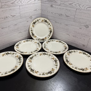 May include: A set of six oval-shaped plates. The plates have a white center with a decorative border of green and brown leaves and a gold rim. The plates are arranged on a black surface.