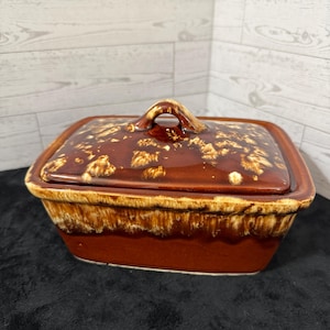 May include: A rectangular ceramic butter dish with a lid, featuring a glossy brown glaze with cream-colored drip patterns along the edges and top. The lid includes a central arched handle, and the base has a smooth, solid brown finish.