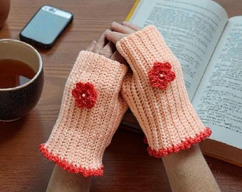 May include: A pair of peach-colored fingerless gloves made of ribbed crochet knit. Each glove features a small, bright red crocheted flower appliqué centered on the back of the hand, with a matching red scalloped crochet trim along the bottom edge.
