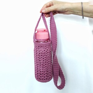 A pink crocheted water bottle holder with a long strap. The holder is made of a delicate, open-weave pattern and is perfect for carrying a water bottle on the go.