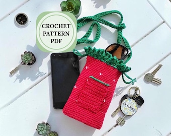 May include: A red crocheted strawberry-shaped phone bag with a green strap and top. The bag has a small pocket and white dots. Sunglasses and keys are inside the bag. The text "CROCHET PATTERN PDF" is in a circle.