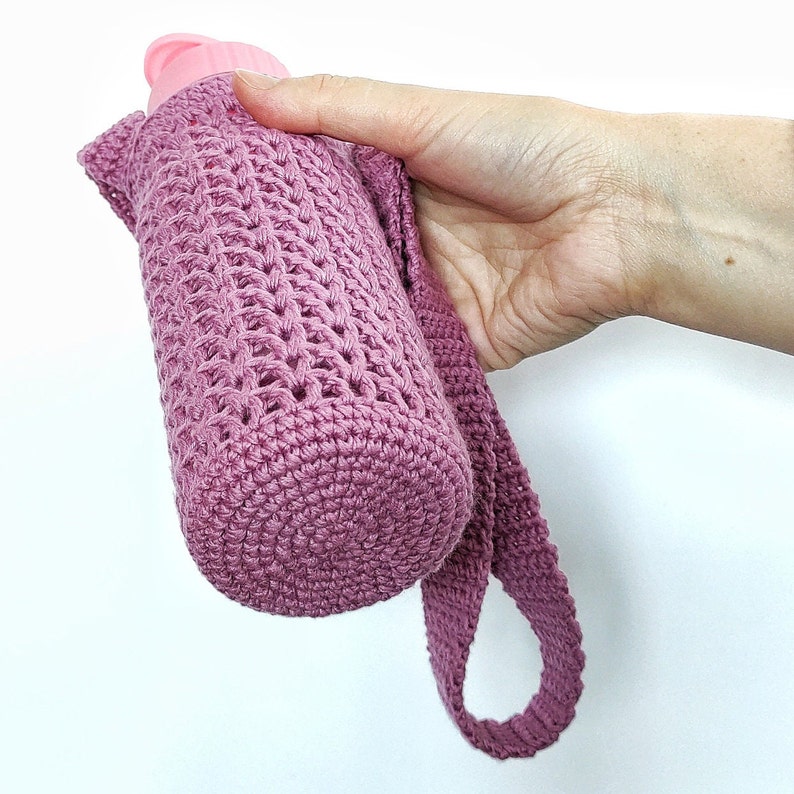 A pink crocheted bottle holder with a strap. The holder is made with a simple stitch pattern and has a round bottom.
