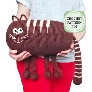 Könnte beinhalten: Eine handgefertigte braune Häkelkatze mit weißen Streifen, Ohren und einem Schwanz. Die Katze hat ein lächelndes Gesicht mit weißen Schnurrhaaren und großen Augen. Der Text "CROCHET PATTERN PDF" befindet sich in einem grünen Kreis.
