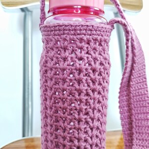 A pink crocheted bottle holder with a long strap. The holder is made with a delicate stitch pattern and is designed to fit a standard water bottle.