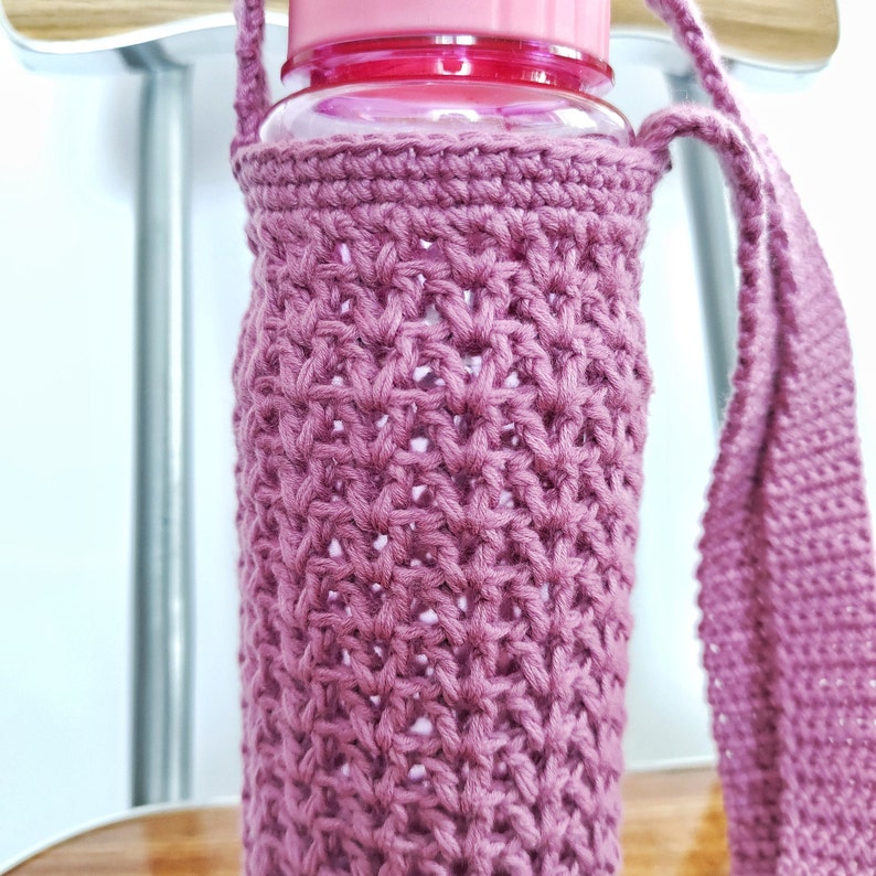 A pink crocheted bottle holder with a long strap. The holder is made with a delicate stitch pattern and is designed to fit a standard water bottle.