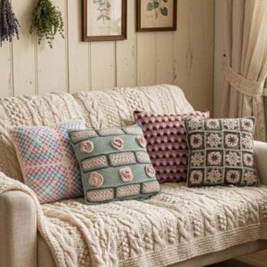 Op de afbeelding: Een crèmekleurige kabelgebreide plaid over een bank, voorzien van vier decoratieve vierkante kussens. De kussens hebben een blauw en roze granny square patroon, een saliegroen ontwerp met verhoogde roze rozetten, een veelkleurig ruitpatroon en een bruin en crème bloemrijk granny square motief.