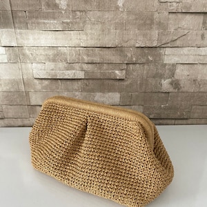 May include: A woven straw clutch bag with a brown color. The bag has a rectangular shape with a rounded top and a clasp closure.