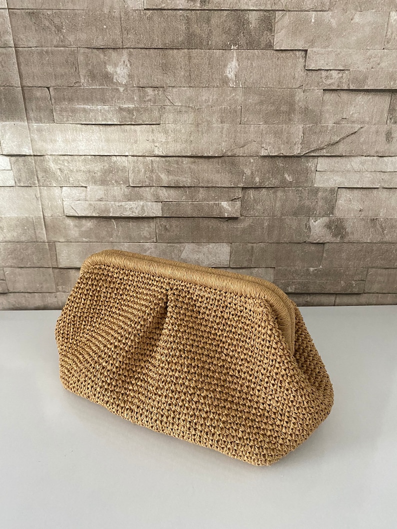 May include: A woven straw clutch bag with a brown color. The bag has a rectangular shape with a rounded top and a clasp closure.
