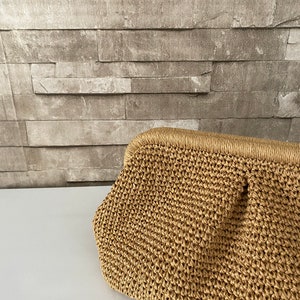 May include: A woven straw clutch bag with a light brown color. The bag has a slightly rounded shape and a smooth texture.
