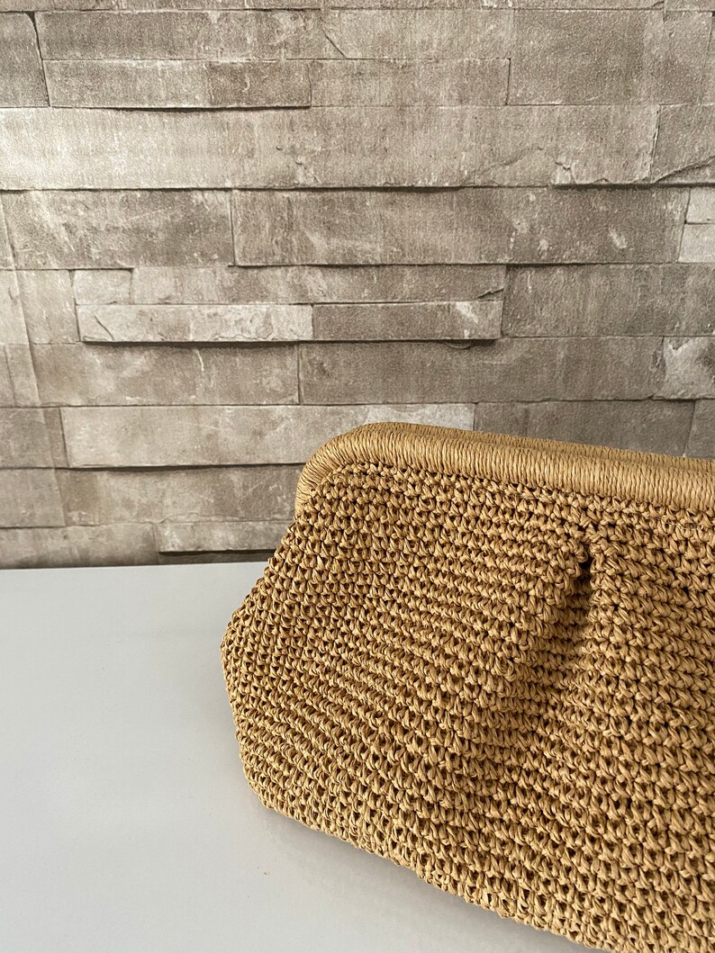 May include: A woven straw clutch bag with a light brown color. The bag has a slightly rounded shape and a smooth texture.