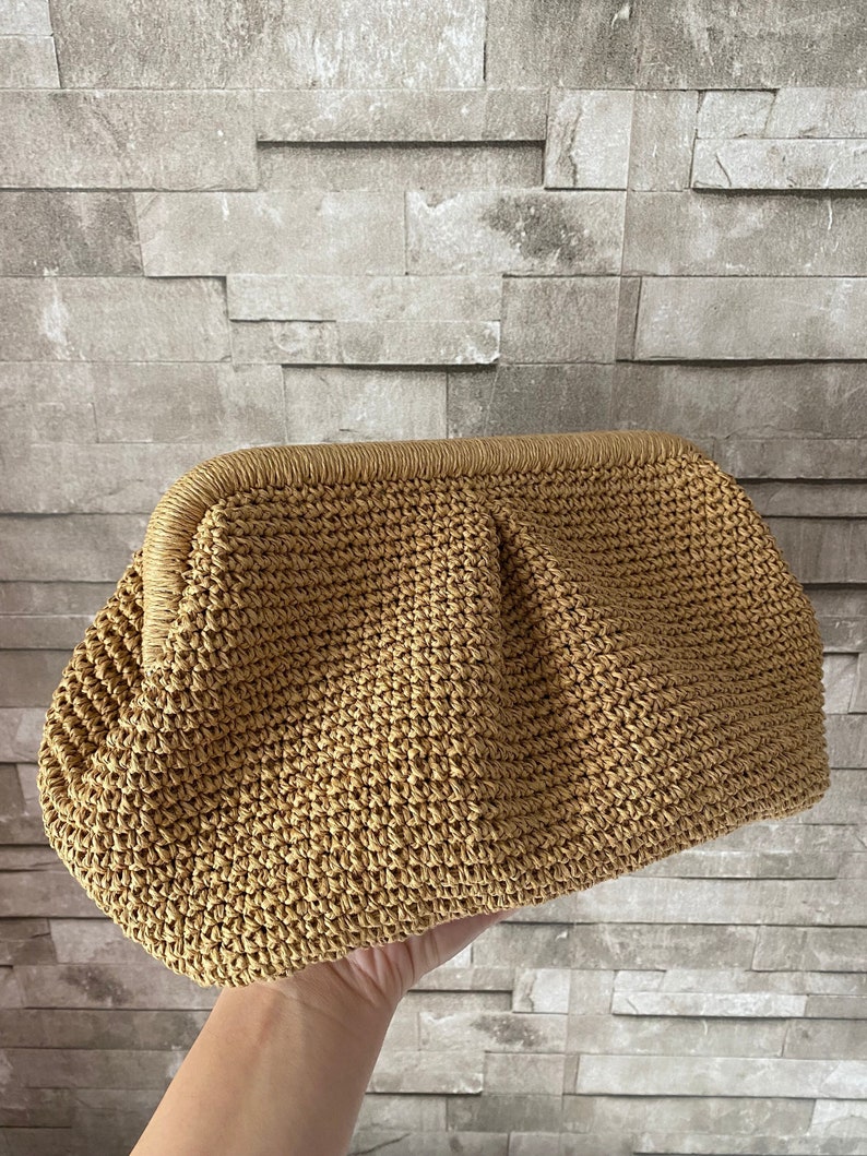 May include: A woven straw clutch bag with a brown color. The bag has a rounded shape and a slightly textured surface.