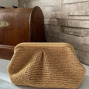 May include: A woven straw clutch bag with a brown color and a slightly rounded shape.