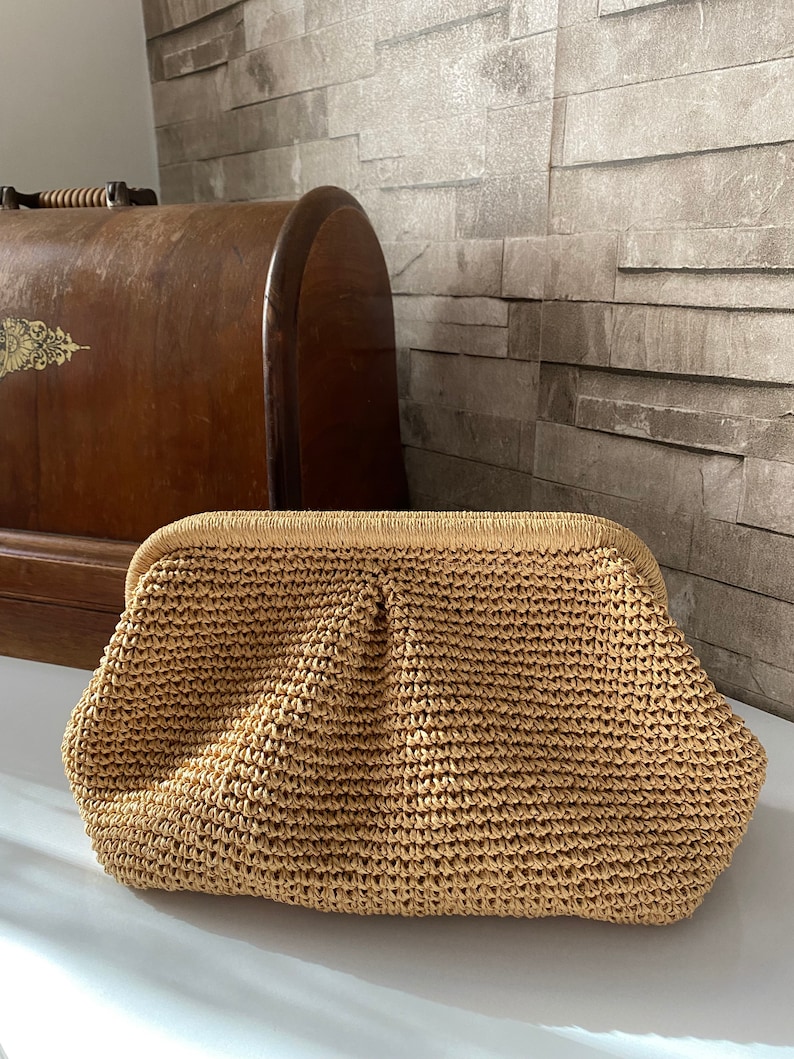 May include: A woven straw clutch bag with a brown color and a slightly rounded shape.